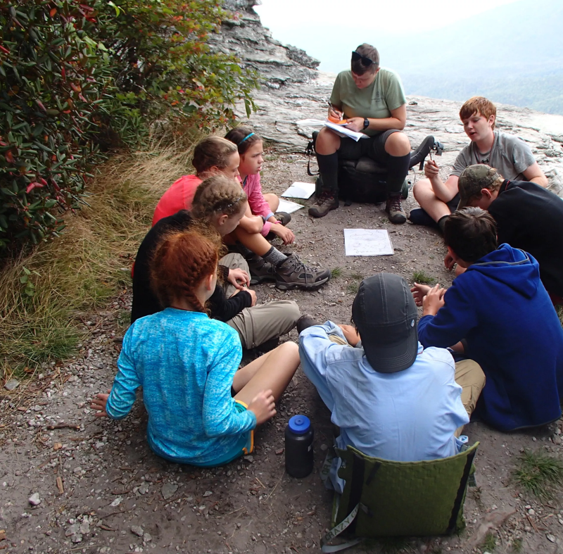 A group of about ten young people are sitting in a circle on a rocky outcrop, seemingly engaged in a lesson or activity. An adult figure is seated slightly apart, taking notes on a clipboard. The setting appears to be outdoors, possibly on a hiking trail or at a scenic overlook, with trees and a distant view visible in the background.